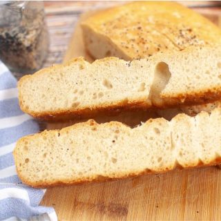 A sliced loaf of sourdough discard focaccia bread on a wooden cutting board, with a striped cloth and a jar of seasoning visible in the background.