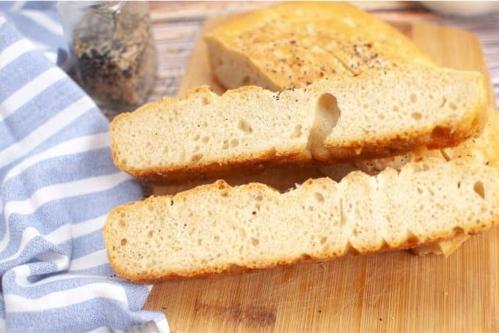 A sliced loaf of sourdough discard focaccia bread on a wooden cutting board, with a striped cloth and a jar of seasoning visible in the background.