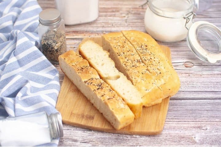 Sliced bread with seasoning sits on a wooden cutting board, surrounded by a striped cloth, glass jars of seasoning, and a container of milk on a wooden surface.