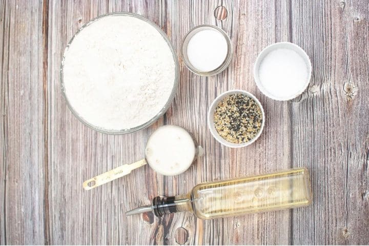 An overhead view of baking ingredients for sourdough discard focaccia recipe on a wooden surface, including flour, sugar, baking powder, seeds, oil in a bottle, a measuring spoon, and a cup with liquid.