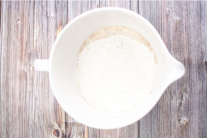 A white mixing bowl containing flour placed on a wooden surface.