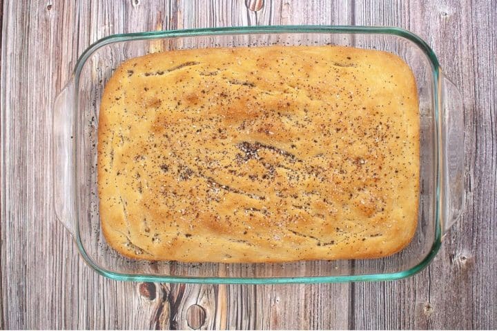 A rectangular loaf of freshly baked sourdough focaccia bread with a golden-brown crust and sprinkled with seeds, sitting in a clear glass baking dish on a wooden surface.
