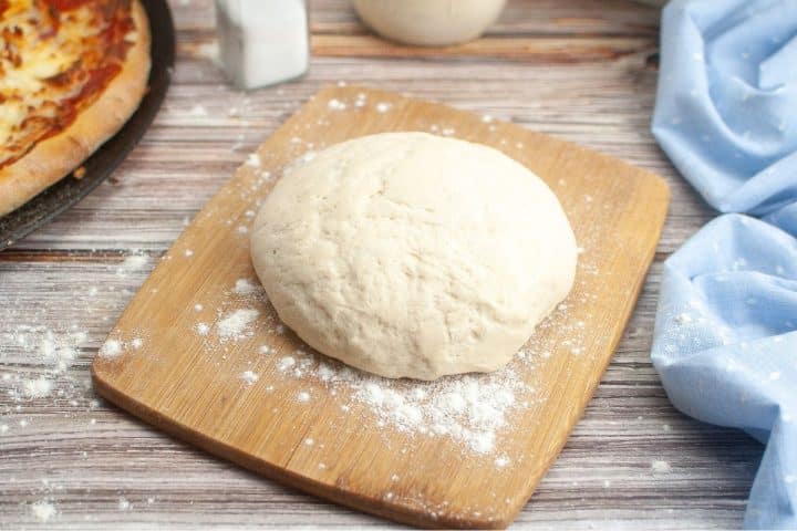 A ball of pizza dough rests on a wooden board, surrounded by a sprinkle of flour, with a pizza in the background and a blue cloth to the side.