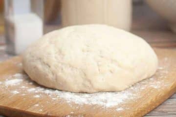 A ball of raw sourdough pizza dough rests on a lightly floured wooden cutting board.