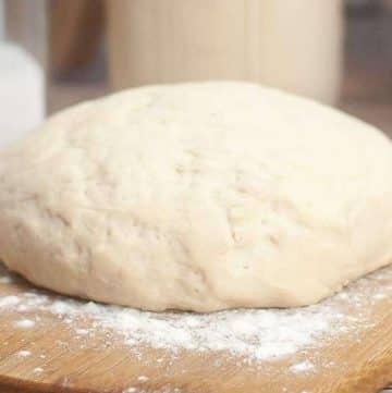 A ball of raw sourdough pizza dough rests on a lightly floured wooden cutting board.