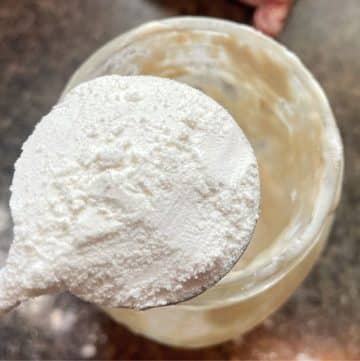 A spoonful of white powder is held over a glass jar on a dark countertop.