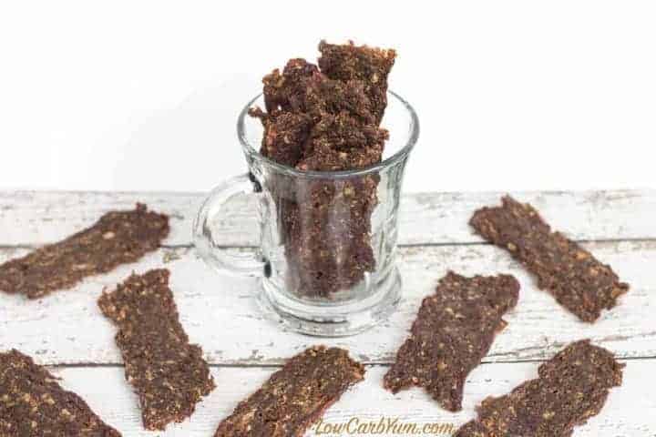 Several pieces of homemade beef jerky are arranged on a white wooden surface, with some pieces standing upright in a clear glass mug—perfect inspiration for your next venison dinner ideas.