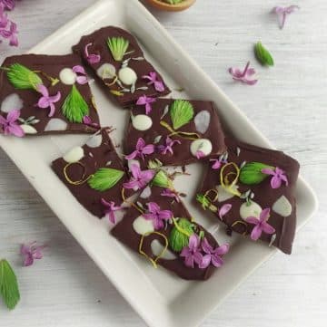 Dark chocolate bark pieces garnished with purple flowers and green leaves on a rectangular white plate, surrounded by scattered flowers and leaves on a light surface.
