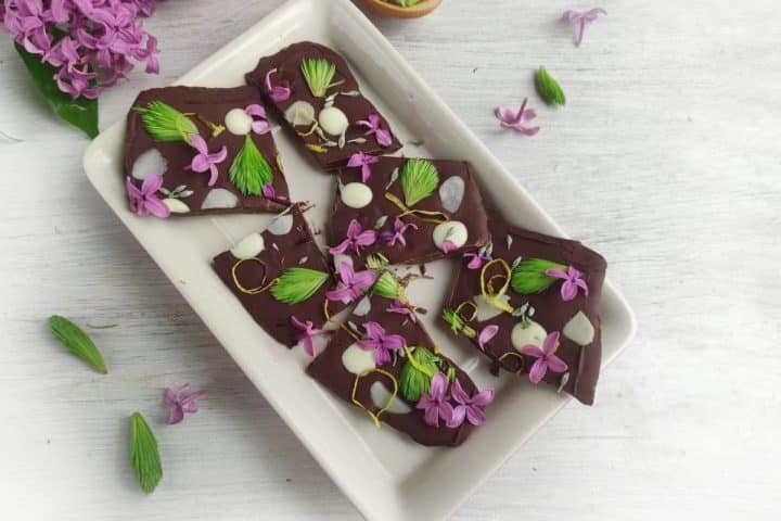 Dark chocolate bark pieces garnished with purple flowers and green leaves on a rectangular white plate, surrounded by scattered flowers and leaves on a light surface.