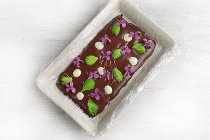 Chocolate cake with purple flowers, white dots, and green leaves on top, set in a rectangular dish on a white surface.