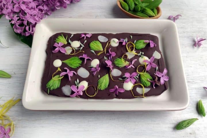 A rectangular dessert with a dark chocolate base is decorated with purple flowers, green leaves, and white dollops, placed on a white rectangular plate.