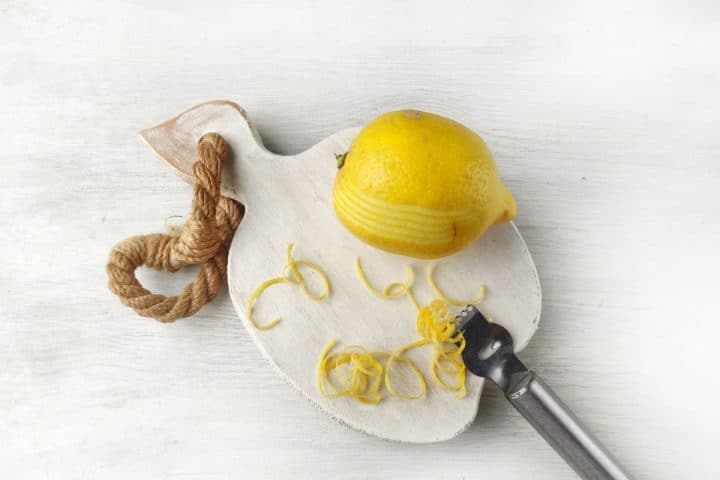 A lemon partially peeled with a zester rests on a white cutting board with a rope handle. Peelings are scattered on the board.