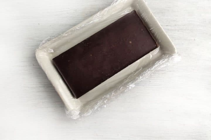 A rectangular block of chocolate wrapped in plastic, placed on a light-colored rectangular tray, viewed from above.