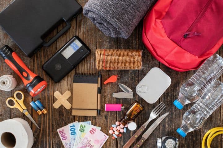Top view of emergency supplies including a first aid kit, flashlight, radio, water bottles, medications, notepad, money, blanket, canned food, and various tools arranged on a wooden surface.