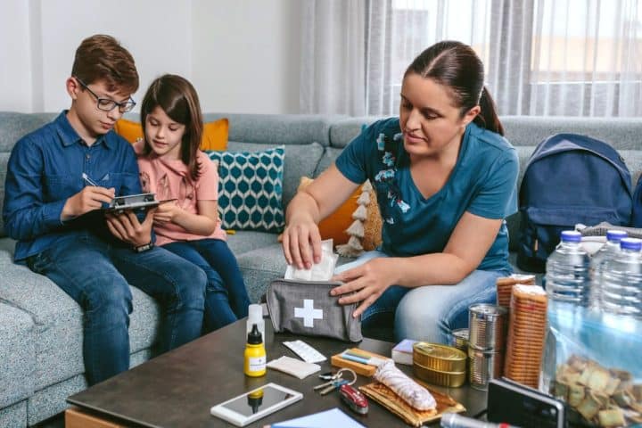A woman packs a first-aid kit while two children look at a clipboard on a couch. Nearby, there are emergency supplies such as water bottles, canned food, and a flashlight.