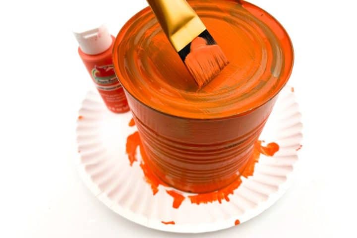 A paintbrush rests on a large tin can covered in orange paint, with a bottle of red acrylic paint nearby on a white paper plate.
