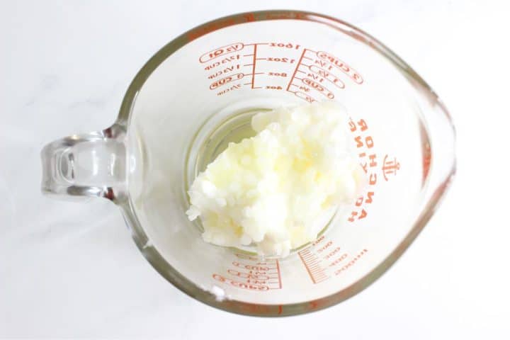 A glass measuring cup filled with solid white coconut oil on a light surface.