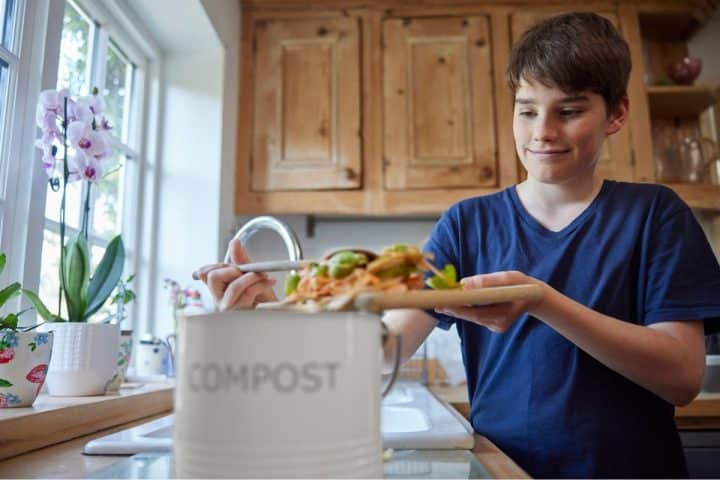 A young person in a kitchen scrapes food scraps from a plate into a compost bin labeled "COMPOST.