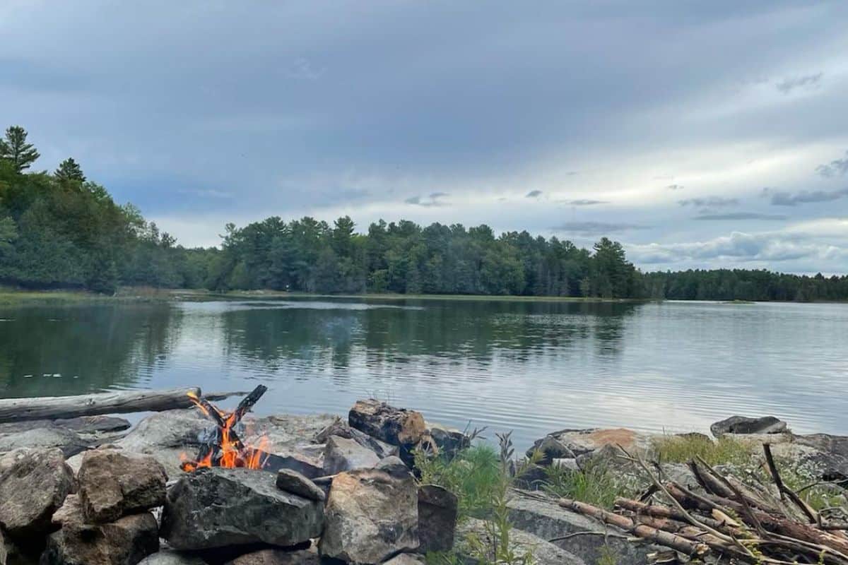 A camsite on a calm lake surrounded by dense trees under a cloudy sky. In the foreground, there is a small campfire tent camping gear, rocks and logs arranged around it.
