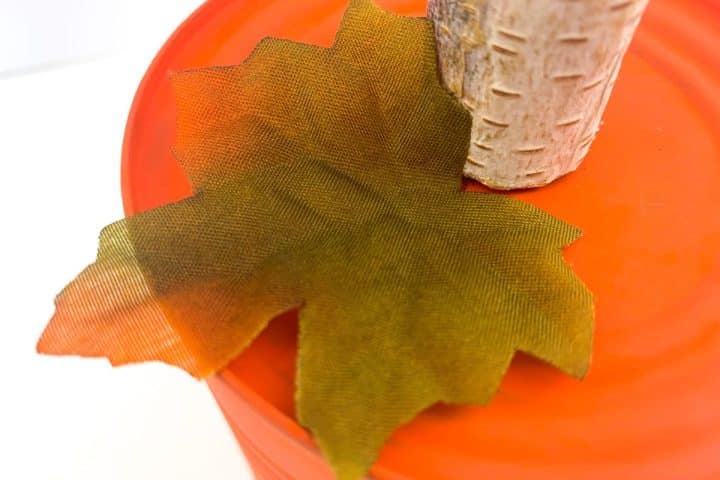 An artificial green leaf next to a faux tree trunk on a red circular surface.