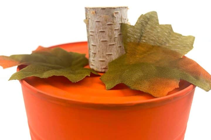 Orange container with a birch-wood stub and two green fabric leaves on top, resembling a pumpkin.