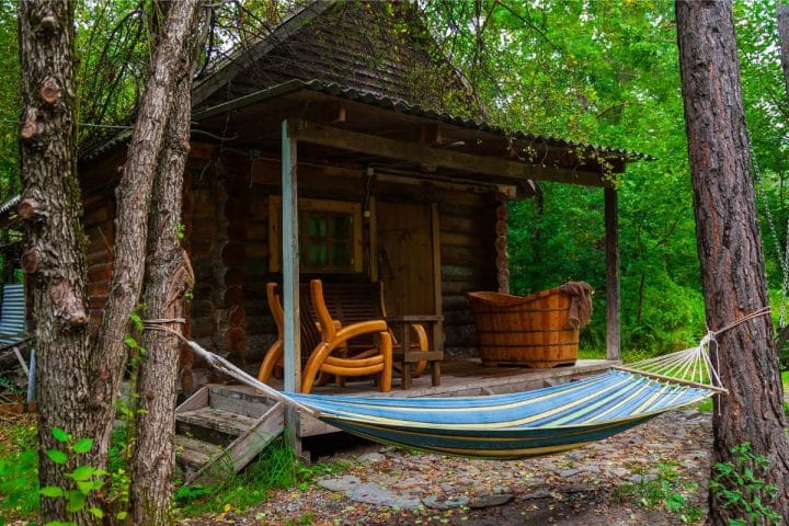 A rustic wooden cabin with a small porch, two chairs, a wooden barrel, and a blue-striped hammock in a lush forest setting.