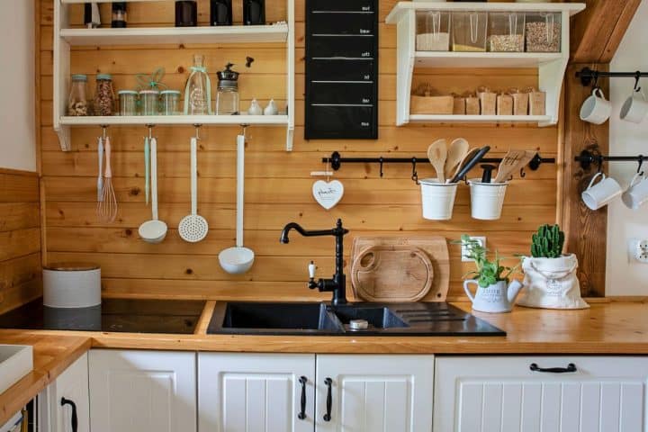 A neatly organized kitchen in a tiny home features wooden shelves with various utensils, a black sink, and spice jars. A cactus plant in a white pot adds charm, while the wood-paneled backsplash and white cabinets below the counter make it functional and stylish, illustrating ideal tiny home storage ideas.