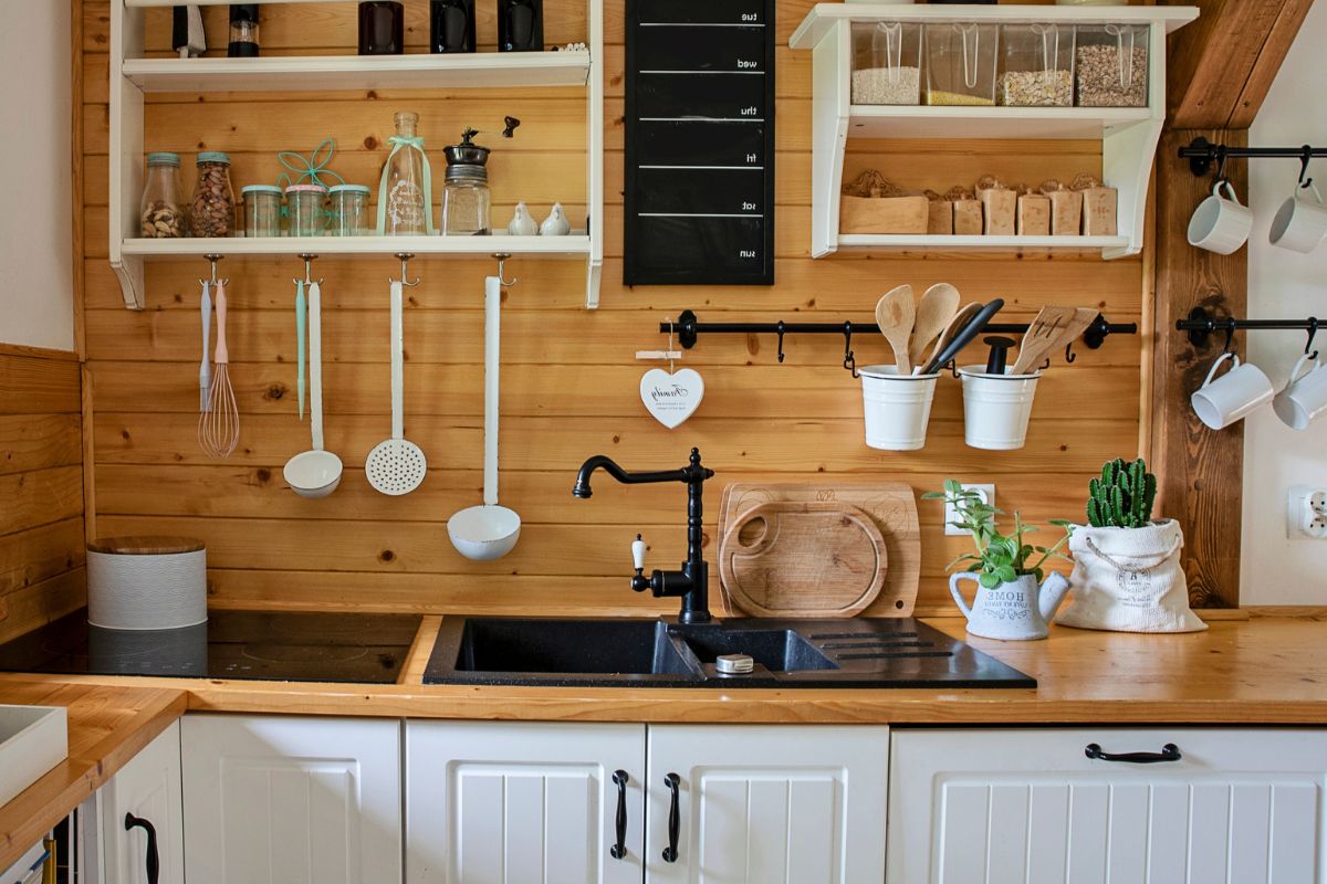 A neatly organized kitchen in a tiny home features wooden shelves with various utensils, a black sink, and spice jars. A cactus plant in a white pot adds charm, while the wood-paneled backsplash and white cabinets below the counter make it functional and stylish, illustrating ideal tiny home storage ideas.