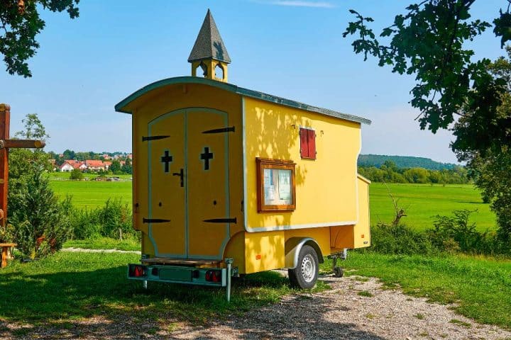 Yellow tiny house on wheels with a steeple, cross-shaped windows, and a landscape background featuring green fields and distant houses.