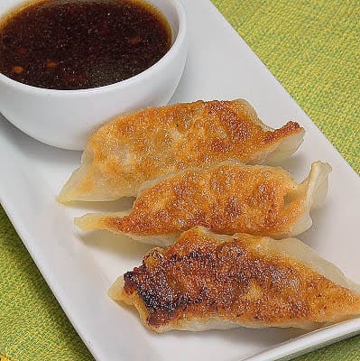 Three pan-fried dumplings are arranged on a rectangular white plate with a small bowl of dipping sauce beside them—perfect for those seeking unique venison dinner ideas.
