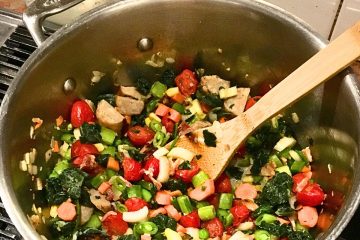 A large pot filled with cooked vegetables including cherry tomatoes, bell peppers, and leafy greens, with a wooden spoon resting inside.