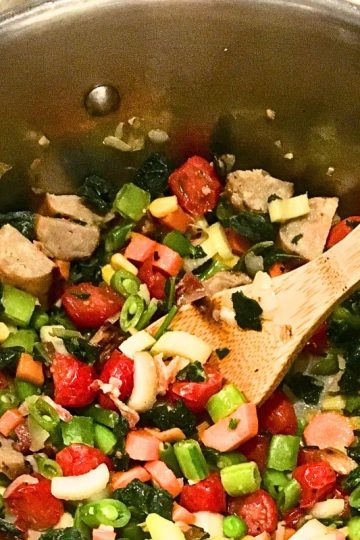 A large pot filled with cooked vegetables including cherry tomatoes, bell peppers, and leafy greens, with a wooden spoon resting inside.