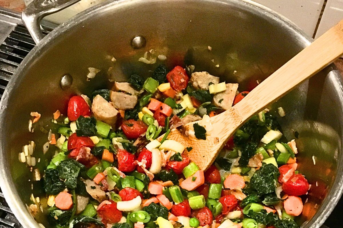 A large pot filled with cooked vegetables including cherry tomatoes, bell peppers, and leafy greens, with a wooden spoon resting inside.