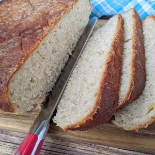 A loaf of whole grain artisan bread is partially sliced on a wooden cutting board with a bread knife; a blue checkered cloth is in the background.