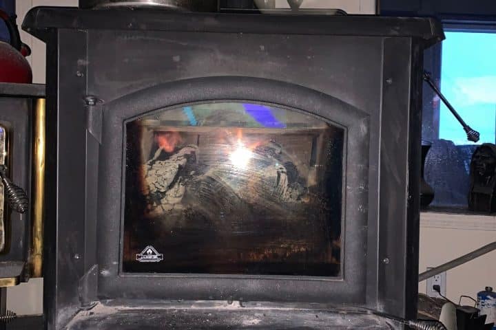 A black wood stove with a glass window, partially obscured by a light reflection, shows the interior and some soot. A red kettle is visible on the left side. A blue-tinted window is in the background.