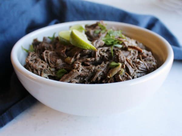 Shredded beef in a white bowl, garnished with lime wedges and fresh herbs, placed on a blue cloth—perfect for those seeking venison dinner ideas with a similar flavorful presentation.