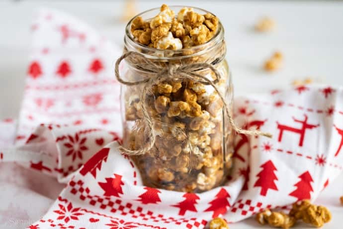 A glass jar filled with caramel popcorn, perfect for homemade food gifts, is tied with twine and placed on a red and white holiday-patterned cloth.