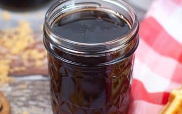 A glass jar filled with pancake syrup on a wooden surface, next to a red and white checkered cloth.