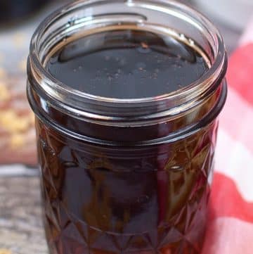 A glass jar filled with pancake syrup on a wooden surface, next to a red and white checkered cloth.