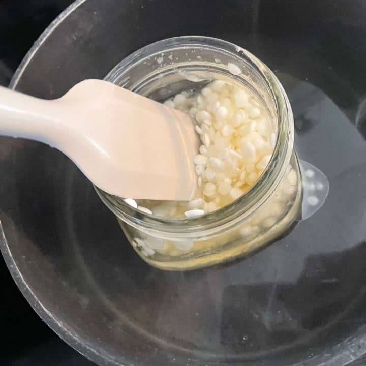 A spoon is being used to stir the beeswax pellets in a jar.