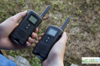two hands each holding a small walkie-talkie outdoors.