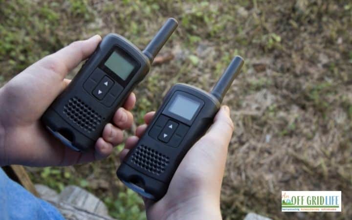 two hands each holding a small walkie-talkie outdoors.