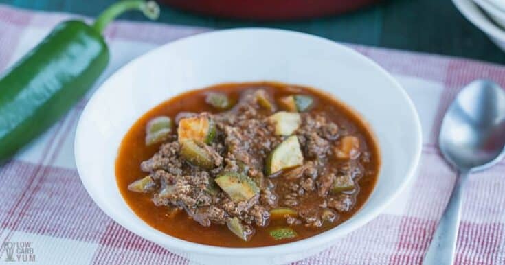 A white bowl filled with chili featuring ground meat and chopped vegetables sits on a red and white checkered cloth next to a spoon and a green pepper—perfect for those seeking venison dinner ideas.