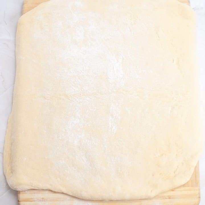 A rolled-out dough on a wooden cutting board, lightly dusted with flour.