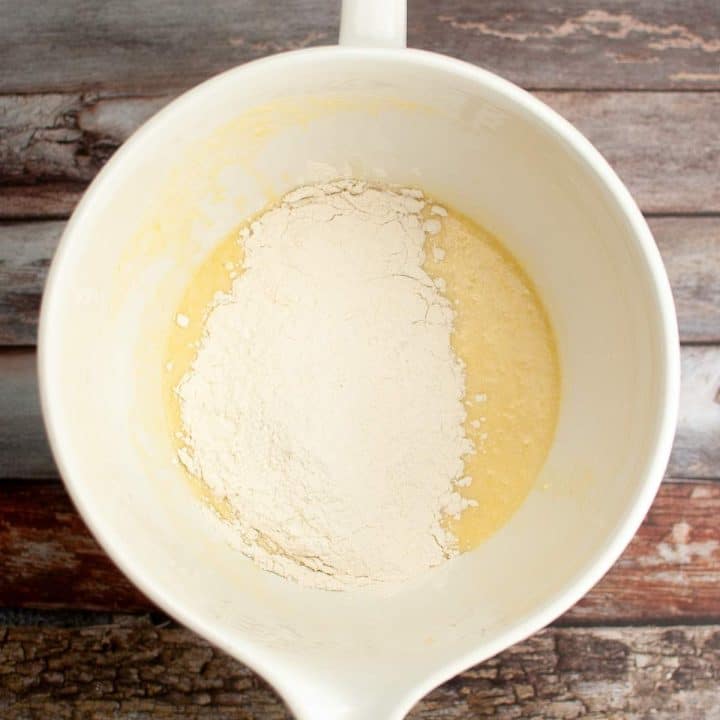 Batter mixture for sourdough donut recipe in a white bowl with dry flour added, ready to be mixed, atop a rustic wooden surface.