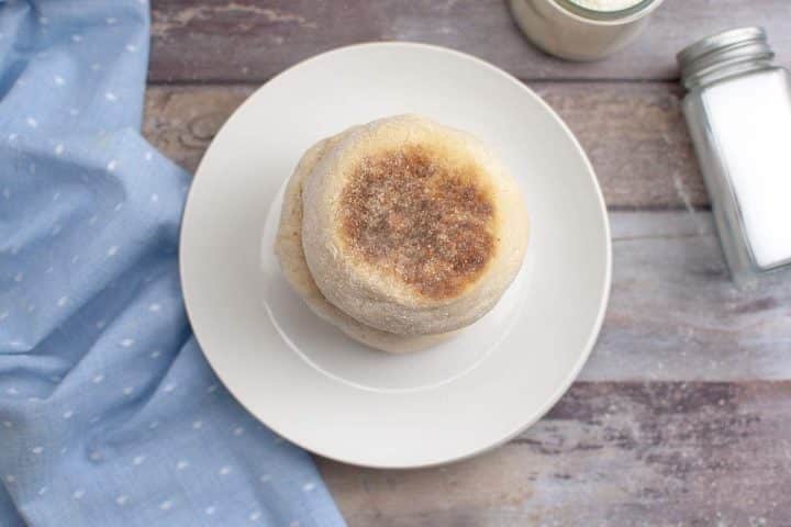 A stack of sourdough english muffins on a white plate, next to a blue cloth and a jar of milk, on a wooden table.