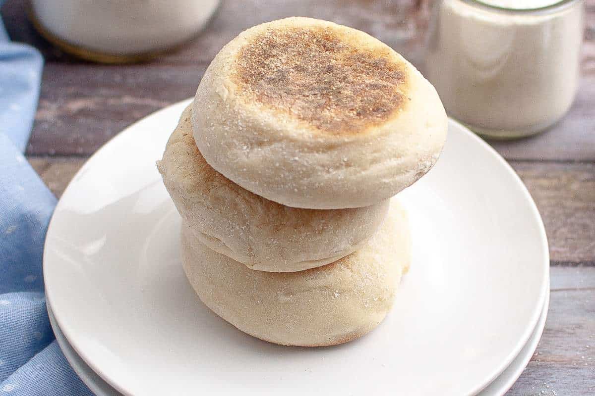 Three freshly baked sourdough english muffins stacked on a white plate, with a rustic wooden background and a glass of milk.
