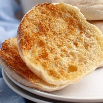 Toasted sourdough english muffin on a white plate with a stack of muffins in the background, set against a blue cloth.