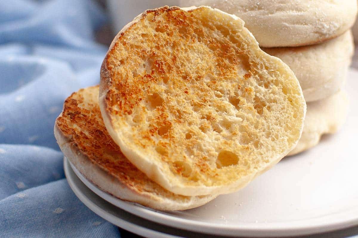 Toasted sourdough english muffin on a white plate with a stack of muffins in the background, set against a blue cloth.