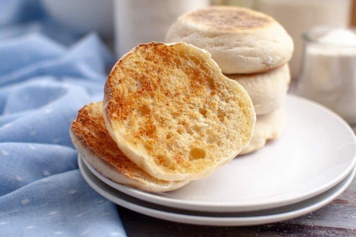 Sourdough English muffins on a plate, one halved and toasted, next to a blue cloth, with a jar of jam in the background.